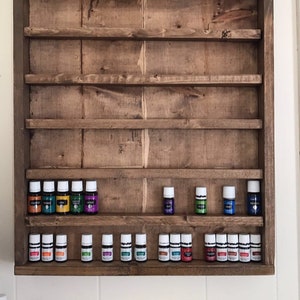 May include: A wooden shelf unit with multiple shelves for storing essential oil bottles. The shelves are stained a dark brown color. The unit is mounted on a white wall. There are 20 essential oil bottles on the shelves, with a variety of colors.