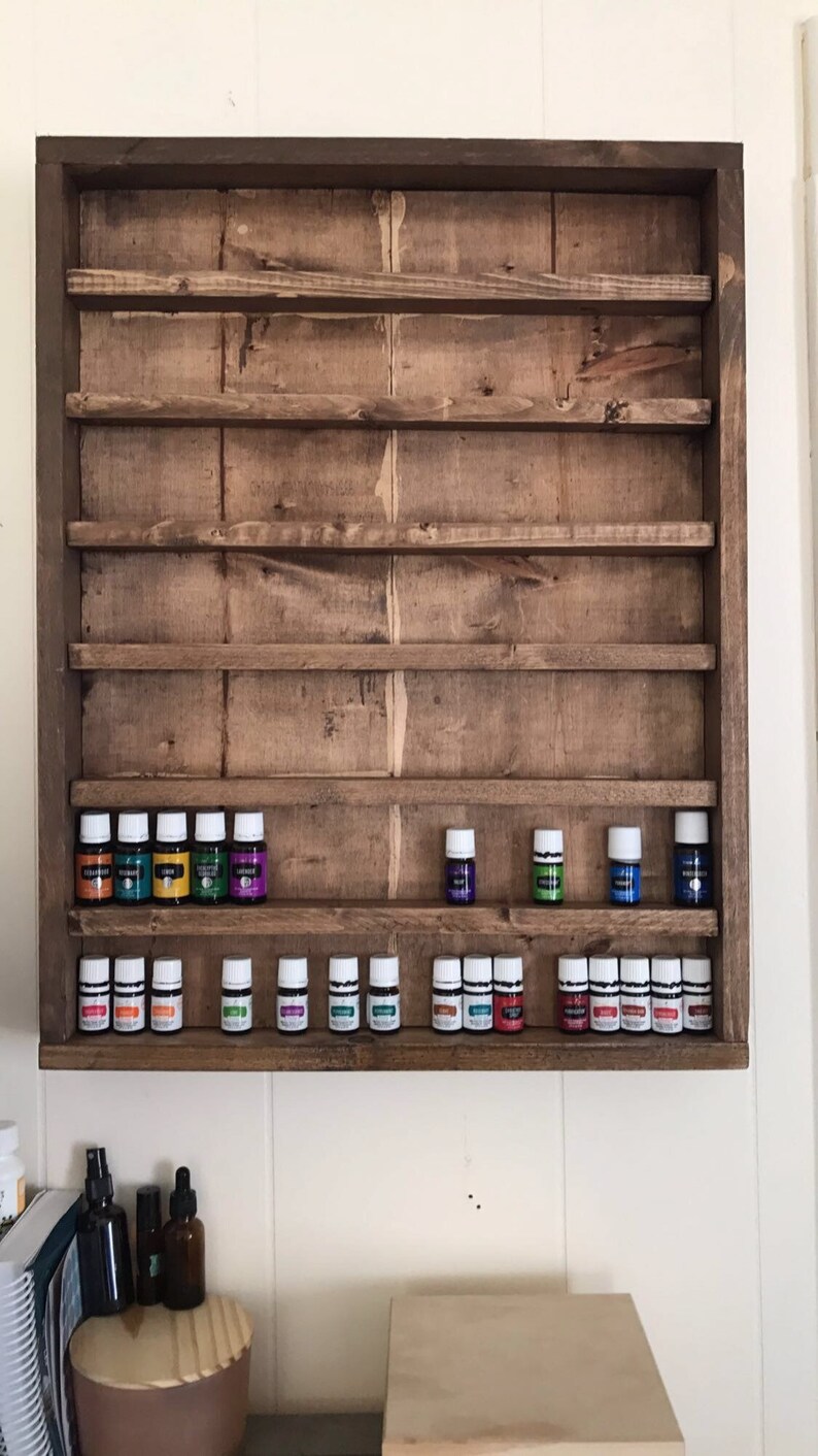 May include: A wooden shelf unit with multiple shelves for storing essential oil bottles. The shelves are stained a dark brown color. The unit is mounted on a white wall. There are 20 essential oil bottles on the shelves, with a variety of colors.