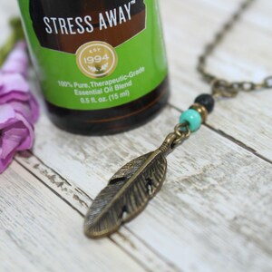 May include: A bronze feather pendant necklace with turquoise and black beads. The necklace is on a white wooden surface with a bottle of essential oil labeled "Stress Away" in the background.