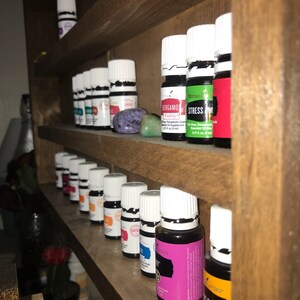May include: A wooden shelf unit with multiple rows of small glass bottles with white caps. The bottles are filled with colorful liquids and have labels with text. The text on the labels includes "Stress Away", "Bergamot", and "Fresh".