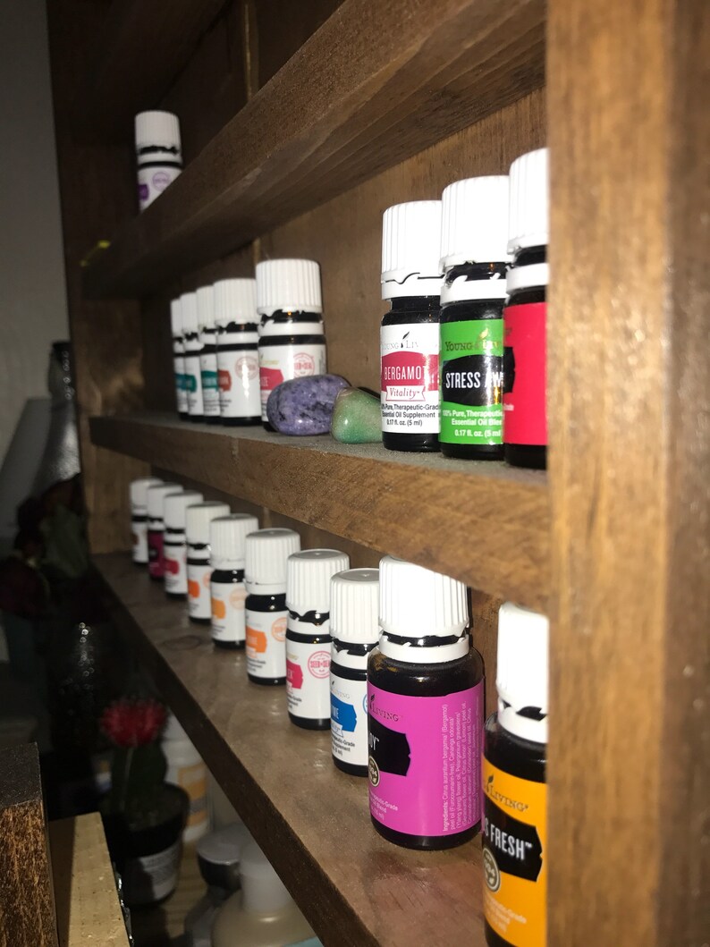 May include: A wooden shelf unit with multiple rows of small glass bottles with white caps. The bottles are filled with colorful liquids and have labels with text. The text on the labels includes "Stress Away", "Bergamot", and "Fresh".