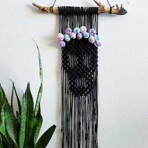 May include: Black macrame skull wall hanging with a crown of pastel-colored flowers. The skull is suspended from a natural wood branch and black cord. A unique, handmade piece of wall decor.