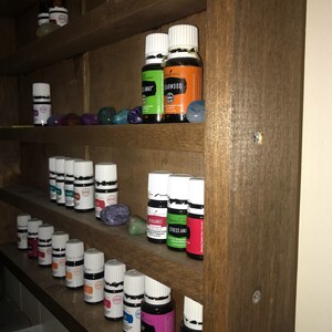 May include: A wooden shelf unit with multiple shelves filled with small glass bottles with white lids. The bottles are labeled with different names, including "Stress Away", "Thieves", "Lemon", "Bergamot", "Cedarwood", and "On Guard".