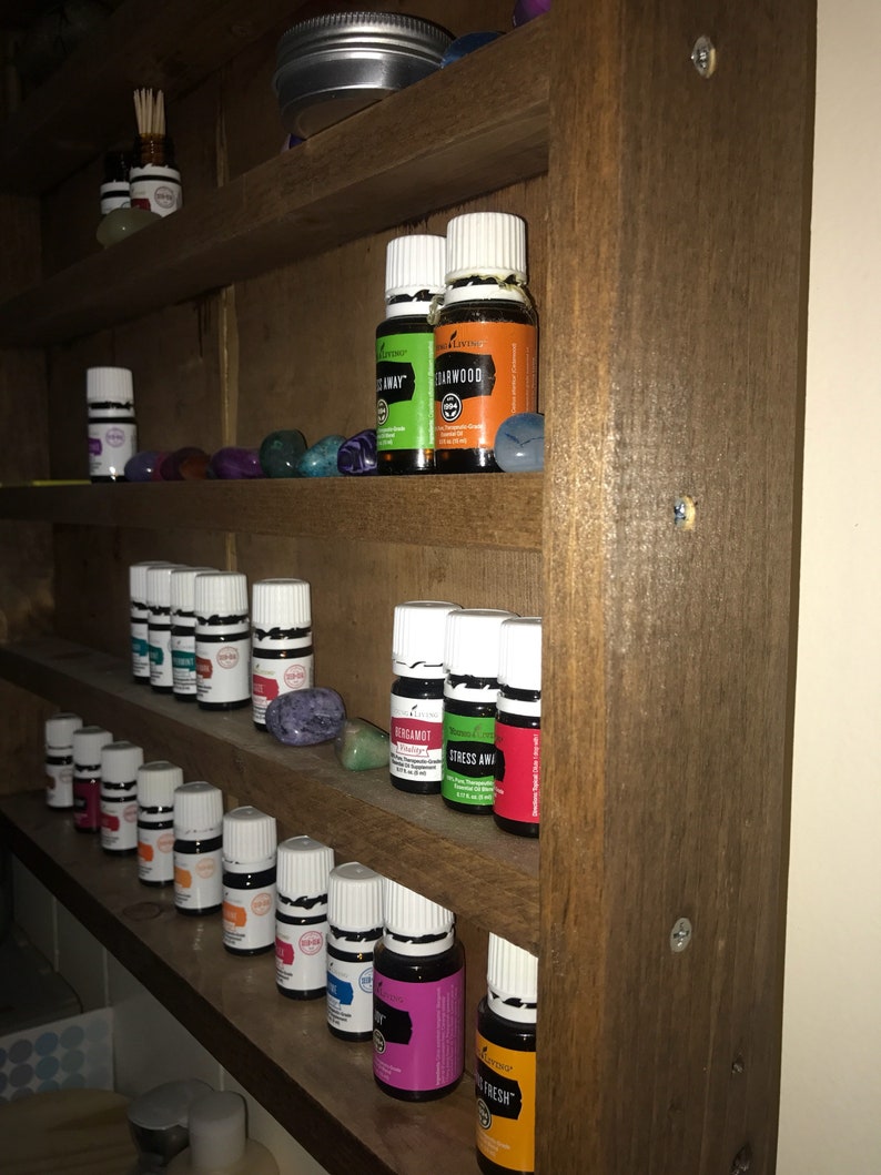 May include: A wooden shelf unit with multiple shelves filled with small glass bottles with white lids. The bottles are labeled with different names, including "Stress Away", "Thieves", "Lemon", "Bergamot", "Cedarwood", and "On Guard".