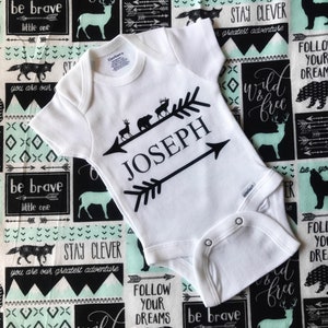 May include: White baby onesie with a black arrow design and the name "JOSEPH" printed on it. The onesie is lying on a black and white fabric with a green and white pattern. The fabric has various phrases printed on it, including "be brave", "stay clever", "wild and free", and "follow your dreams".
