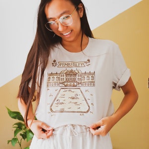 May include: Beige t-shirt with a brown illustration of a stately home and the word "PEMBERLEY" above it. The illustration includes a pond with swans. The shirt is being held up by a person wearing glasses.