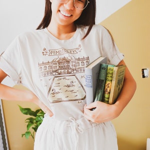 May include: Beige t-shirt with a brown illustration of a building and the word "PEMBERLEY". The person is holding three books. The person is wearing glasses and light-colored pants. The background is a yellow wall.