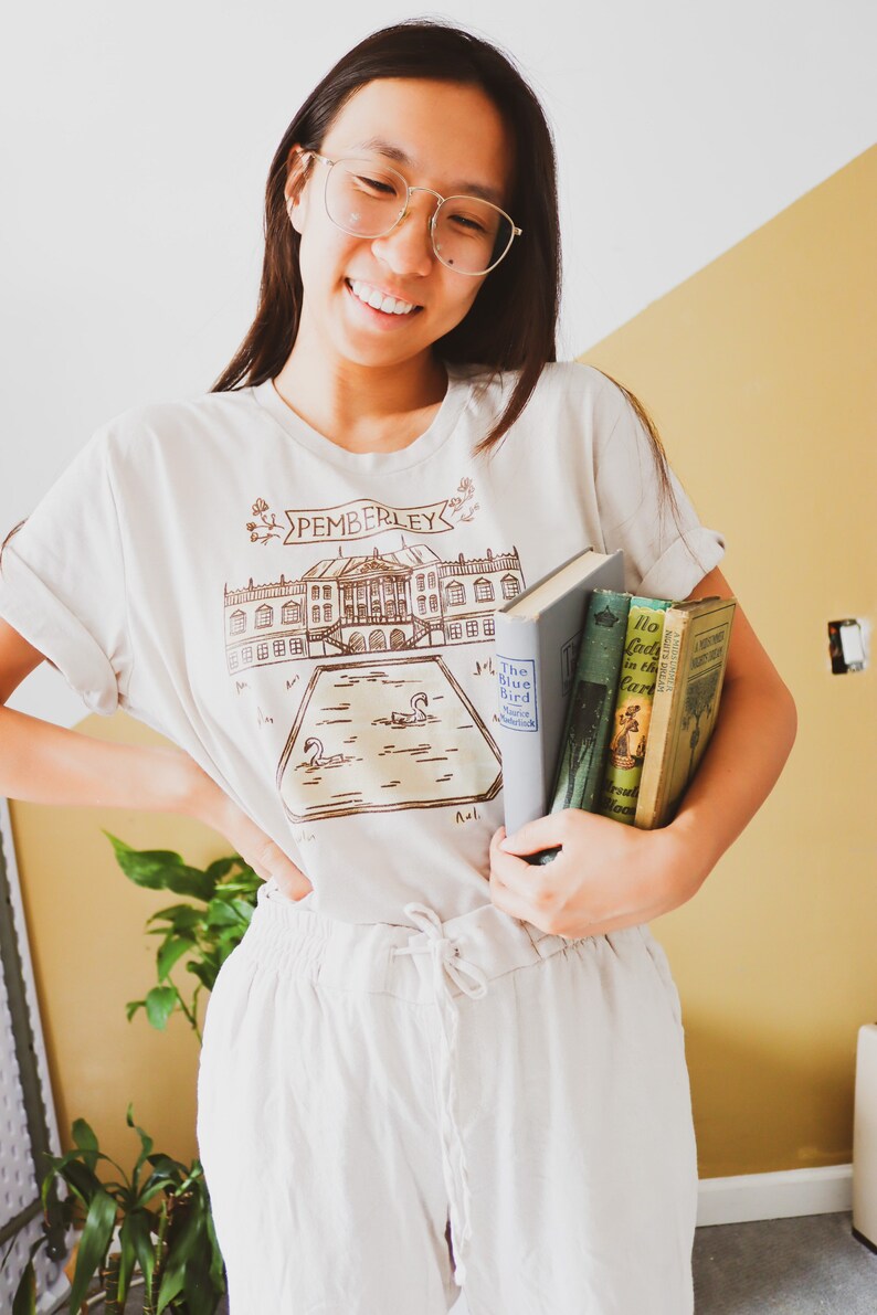 May include: Beige t-shirt with a brown illustration of a building and the word "PEMBERLEY". The person is holding three books. The person is wearing glasses and light-colored pants. The background is a yellow wall.