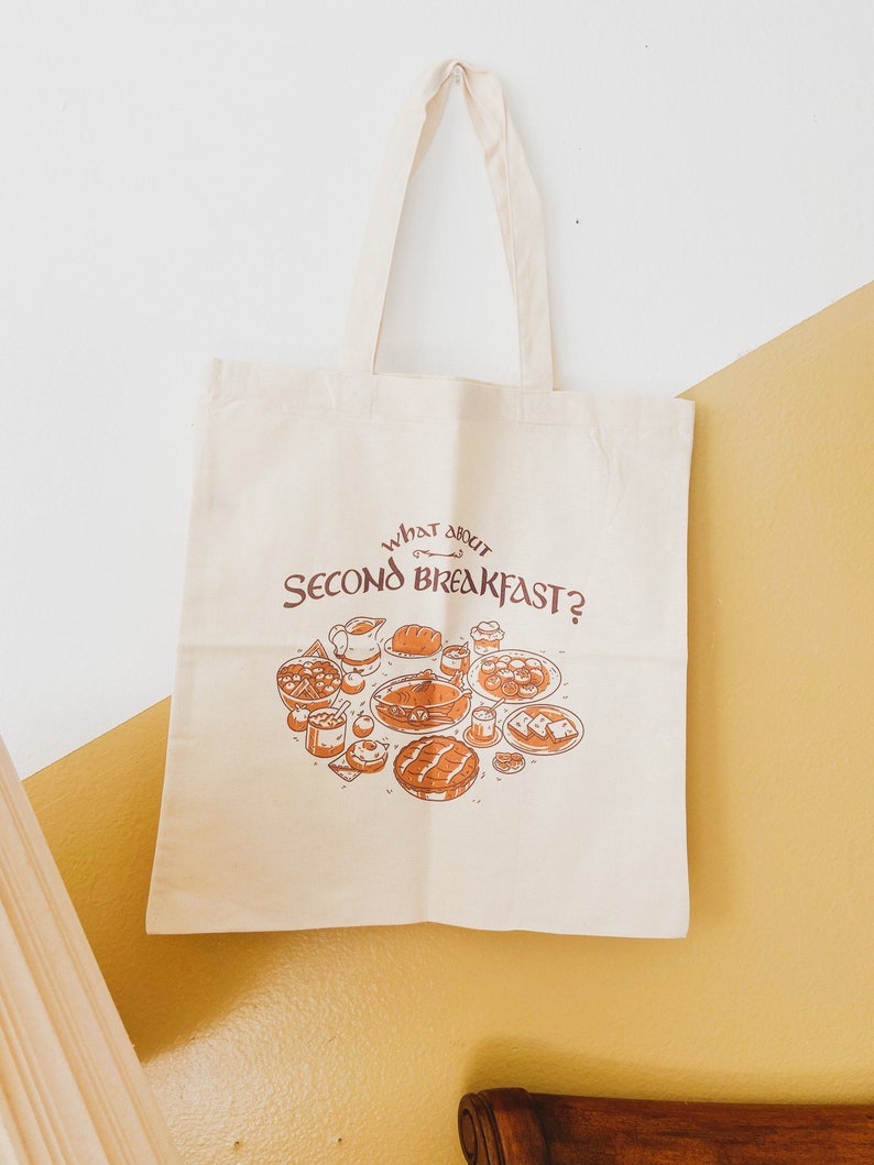 May include: Cream-colored tote bag with the text "WHAT ABOUT SECOND BREAKFAST?" and a brown illustration of various breakfast foods. The bag has two handles and is hanging on a white and yellow wall.