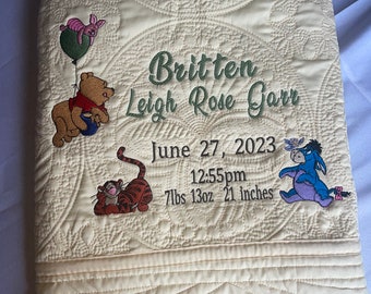 May include: A cream-coloured quilted baby blanket with Winnie the Pooh and friends embroidered on it. The blanket is personalised with the name "Britten Leigh Rose Garr", the date "June 27, 2023", the time "12:55pm", and the baby's weight and length: "7lbs 13oz 21 inches".
