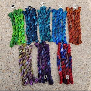 May include: An overhead shot shows eight bundles of colourful embroidery floss. The floss colours include shades of green, blue, orange, purple, and red. Each bundle is labelled with a letter from A to H.