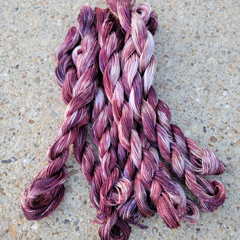 K&ouml;nnte beinhalten: Ein B&uuml;ndel mit sechs Kn&auml;uel Stickgarn in den Farben Burgunderrot, Rosa und Wei&szlig;. Das Garn ist gedreht und hat ein mehrfarbiges Muster.