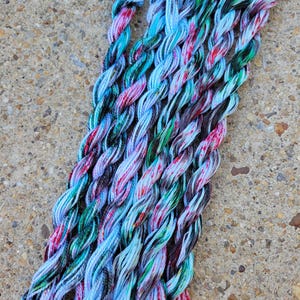 Puede incluir: Hilo de bordar trenzado en tonos de azul, verde, rosa y negro. El hilo está fuertemente torcido, creando una apariencia texturizada. Los hilos de colores están dispuestos en un paquete ordenado, ideal para varios proyectos de manualidades.