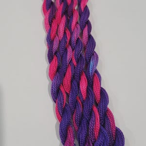 May include: A length of braided embroidery thread in purple and pink hues. The thread is tightly twisted, giving it a textured look. The colours blend, creating a gradient effect. Suitable for embroidery or other craft projects.