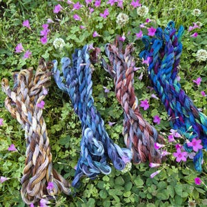 May include: Four bundles of colorful yarn in shades of brown, blue, and purple, arranged on a bed of green clover and small pink flowers. The yarn appears to be hand-dyed, with variations in color throughout each bundle.