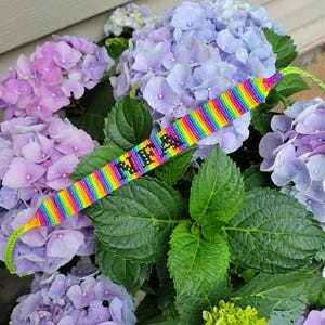 May include: A handmade friendship bracelet with a rainbow striped pattern and the name "MIA" in black letters. The bracelet is displayed on a bed of purple and pink hydrangeas and green leaves.