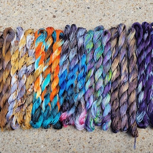 May include: A collection of 28 skeins of hand-dyed yarn in various colors, including shades of brown, blue, green, purple, and orange. The yarn is arranged in rows, with each skein tied together.
