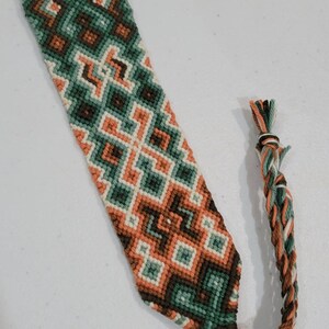 May include: Handmade friendship bracelet featuring a geometric pattern in shades of green, orange, brown, and cream. The woven design includes diamond and zig-zag shapes. The bracelet has a loop at one end and a braided tassel at the other.