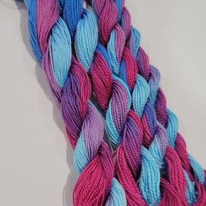 May include: Braided skeins of embroidery floss in shades of blue, pink, and purple. The floss is tightly twisted, creating a textured appearance. The colours blend together, creating a gradient effect. Ideal for embroidery, cross-stitch, or other needlecraft projects.