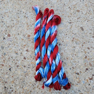 Puede incluir: Cuatro ovillos de hilo en combinaciones de colores rojo, blanco y azul. El hilo está retorcido y tiene un efecto marmoleado.