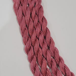 May include: Close-up of several skeins of pink embroidery thread, tightly twisted and arranged in parallel. The thread has a slightly textured appearance, and the colour is a muted, dusty rose. The background is a plain white surface.