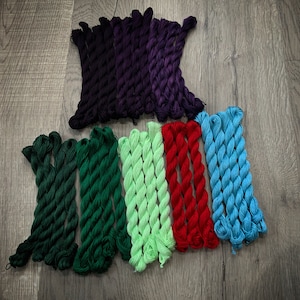 May include: Assortment of embroidery floss in various colors, including green, red, blue, and purple. The floss is wound into skeins and arranged in a row.