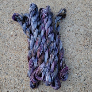 K&ouml;nnte beinhalten: Ein B&uuml;ndel aus violetten und blauen Stickgarn-Kn&auml;ueln. Die Kn&auml;uel sind miteinander verdreht und haben ein buntes Farbmuster.