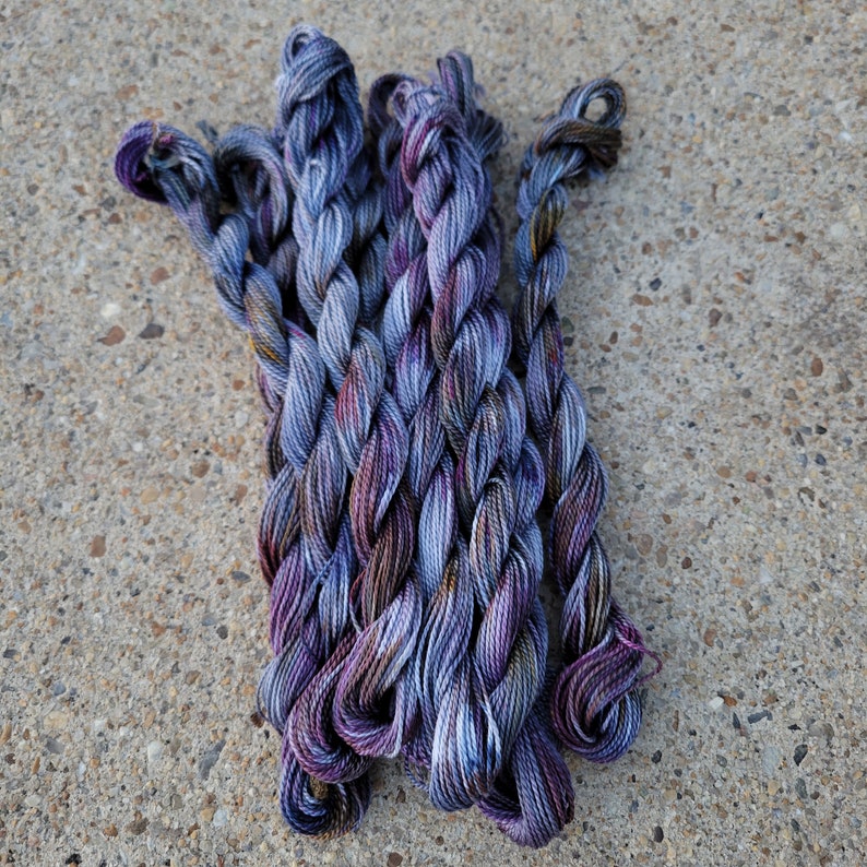K&ouml;nnte beinhalten: Ein B&uuml;ndel aus violetten und blauen Stickgarn-Kn&auml;ueln. Die Kn&auml;uel sind miteinander verdreht und haben ein buntes Farbmuster.