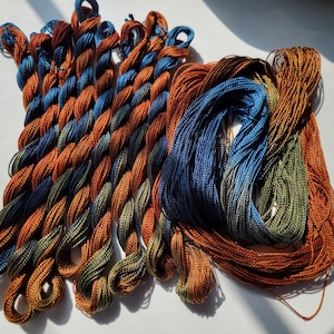 May include: A collection of embroidery floss skeins featuring a variegated color palette of deep blue, burnt orange, and olive green. The threads are presented in both twisted, looped bundles and loose, untwisted hanks of shiny fiber.