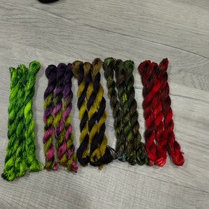 May include: Eight skeins of embroidery floss in various colours, including green, purple, black, and red. The floss is arranged in a row on a light wood surface.