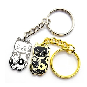 May include: Two lucky cat keyrings. One is silver and white, the other is gold and black. Both feature a chain and a keyring. The cats are decorated with floral patterns and have a raised paw.