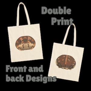 May include: Two off-white canvas tote bags featuring a brown, round frog illustration. The left bag shows the frog's face with a grumpy expression, while the right bag displays the frog's back with a vertical stripe. Text reads Double Print.