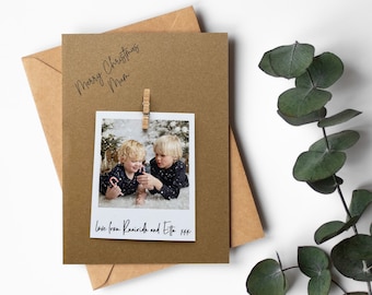 May include: A Christmas card with a kraft paper background and envelope. The card features a photo of two children and the text "Merry Christmas Mum" and "Love from Ruairidh and Etta". A wooden clip holds the photo in place.