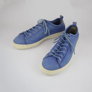 Puede incluir: Un par de zapatillas azules con cordones y suelas blancas. Los zapatos están hechos de un material suave similar al ante. Las zapatillas son de color azul claro y tienen un estilo casual.