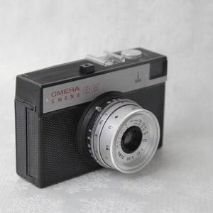 May include: A black and silver 35mm film camera with the text "СМЕНА 8M" on the front. The camera has a silver lens with a black ring around it.