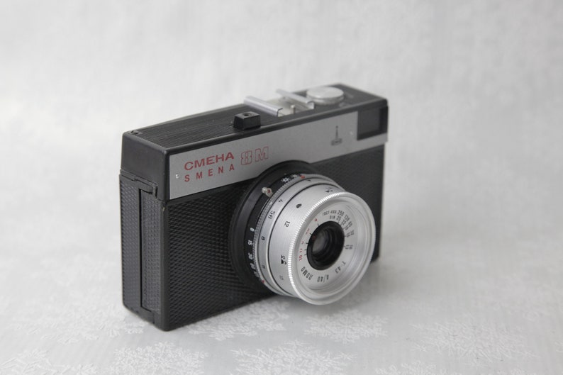 May include: A black and silver 35mm film camera with the text "СМЕНА 8M" on the front. The camera has a silver lens with a black ring around it.