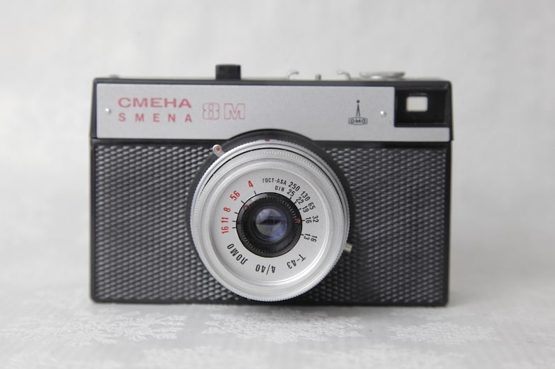 May include: A black and silver 35mm film camera with the text "СМЕНА SMENA 8M" on the top. The camera has a silver lens with a red and white aperture ring.