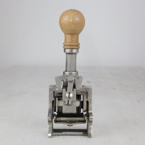 May include: A silver metal number stamp with a wooden handle. The stamp has a dial with numbers 1, 2, 3, and 4.