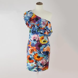 Op de afbeelding: Een jurk met één schouder en een levendige bloemenprint. De jurk heeft een getailleerd silhouet en een gerimpelde halslijn. Het bloemenpatroon bevat blauwe, roze, oranje en gele bloemen tegen een paarse en blauwe achtergrond. De jurk wordt op een mannequin getoond.