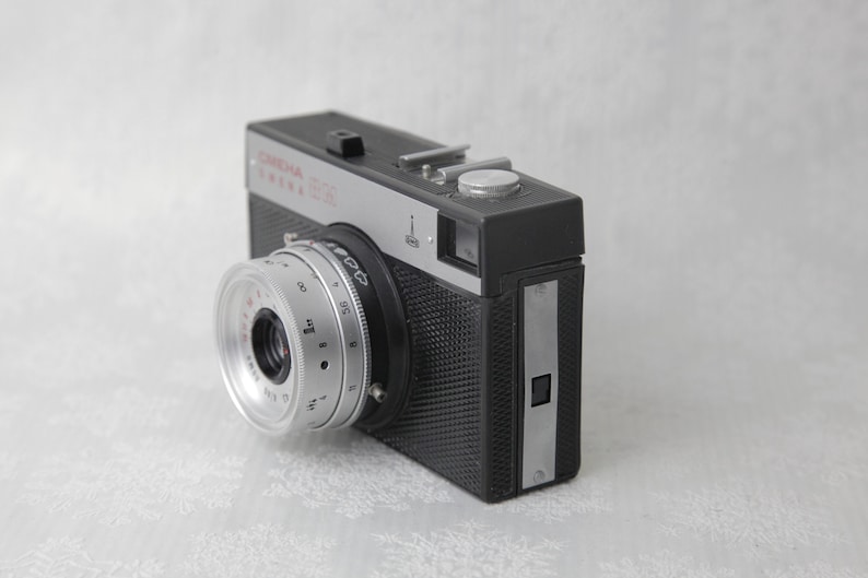 May include: A black and silver 35mm film camera with a silver lens and a black textured body. The camera has a silver nameplate that reads "Corona" and "Corona 35S".