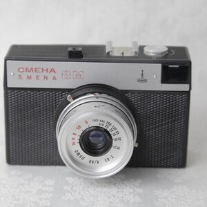 May include: A black and silver 35mm film camera with the text "СМЕНА SMENA 8M" on the top. The camera has a silver lens with a black ring and red markings.