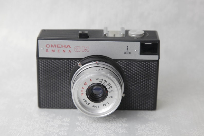 May include: A black and silver 35mm film camera with the text "СМЕНА SMENA 8M" on the top. The camera has a silver lens with a black ring and red markings.