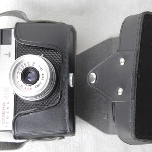 May include: A black and silver 35mm film camera with a leather case. The camera has a lens with a silver ring and a black leather strap. The camera is labeled "Smena" and "OMEHA" in red letters.