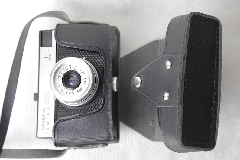 May include: A black and silver 35mm film camera with a leather case. The camera has a lens with a silver ring and a black leather strap. The camera is labeled "Smena" and "OMEHA" in red letters.