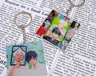 May include: Two acrylic keychains with anime-style illustrations. One features two figures in a frame, the other has a collage of images. Both have silver key rings and chains.