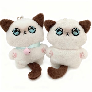 Può includere: Due portachiavi a forma di gatto di peluche. I gatti hanno pelo bianco, orecchie e code marroni e grandi occhi blu. Un gatto indossa un collare azzurro con una campanella rosa. Le zampe hanno impronte rosa. I portachiavi sono progettati per essere carini e coccolosi.