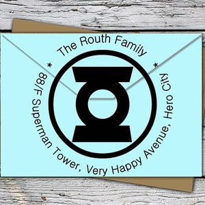 Pode incluir: Um envelope azul claro com o texto "The Routh Family" e um emblema circular preto. O endereço inclui "88/F Superman Tower, Very Happy Avenue, Hero City."