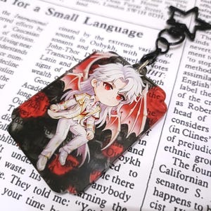 May include: A rectangular pendant featuring an anime-style character with white hair, red eyes, and red wings. The character is dressed in a white suit with gold accents. The background is black with red floral accents and text.