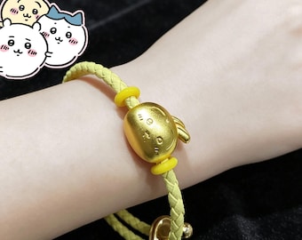 May include: A braided light yellow cord bracelet featuring a central gold-toned charm shaped like a cartoon rabbit head, flanked by two small yellow rings, and finished with a gold-toned metal clasp at the end of the woven cord structure.