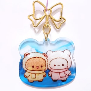 May include: A clear acrylic charm featuring two cartoon bears in winter attire, set against a blue and white winter scene. The charm has a gold-colored bow and clasp for hanging.